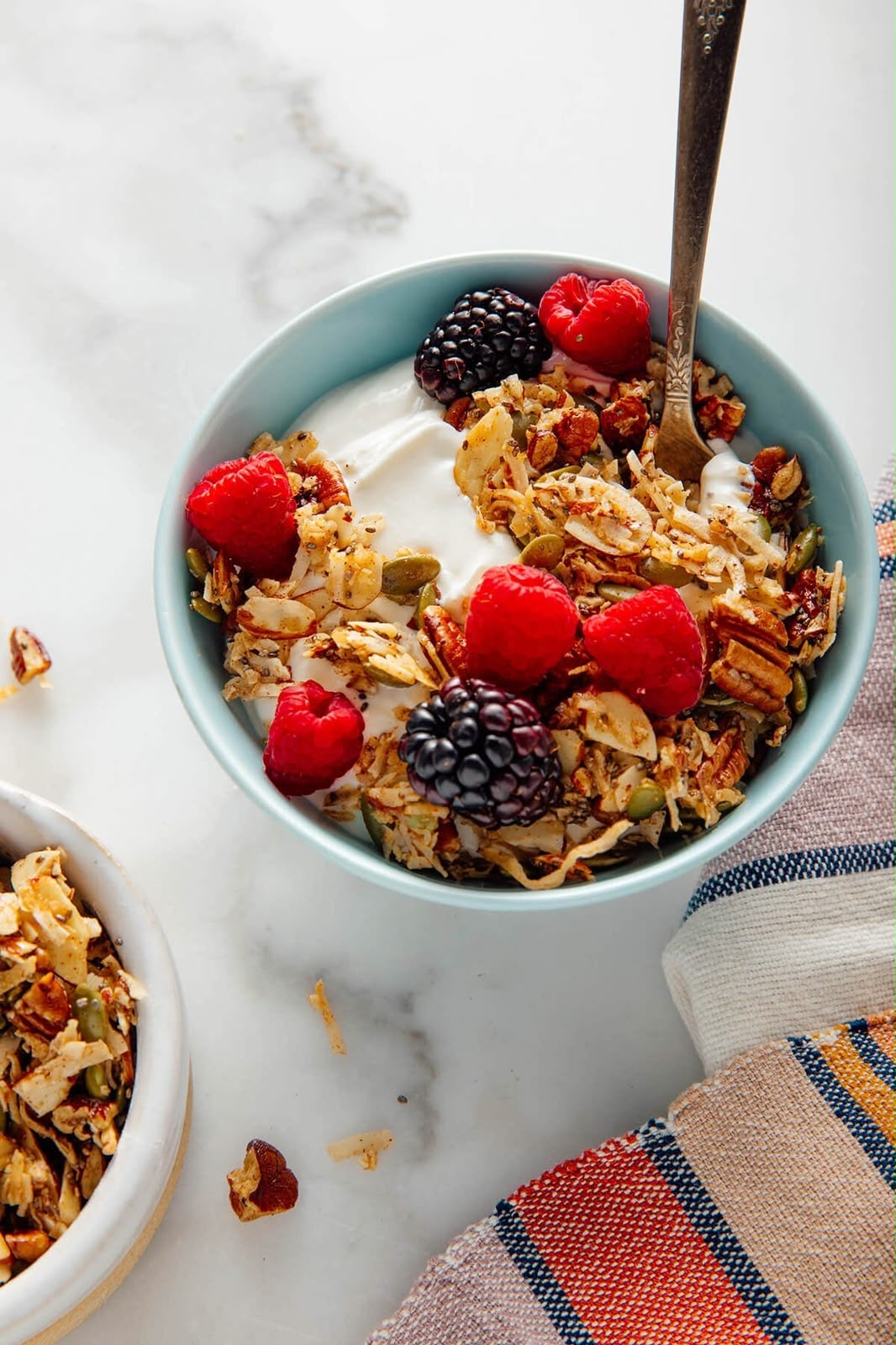 A light blue bowl filled with granola, fresh raspberries, a blackberry, and a dollop of yogurt, with a spoon resting inside. A smaller bowl of granola and a striped cloth napkin are visible on a white marble surface.