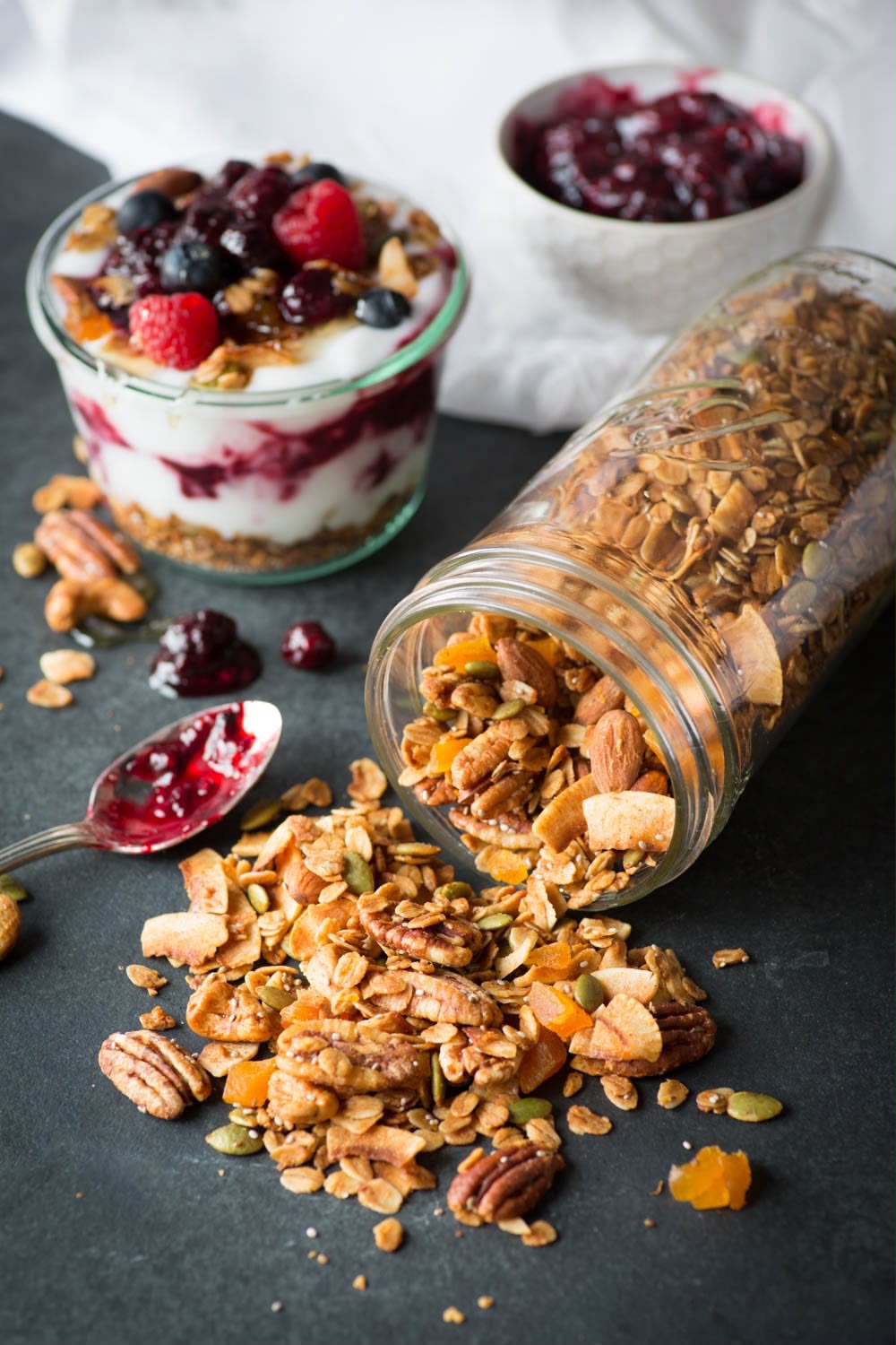 Granola mix spilled from a clear glass jar on a dark surface, with a glass cup of yogurt topped with berries and a small bowl of berry compote in the background.