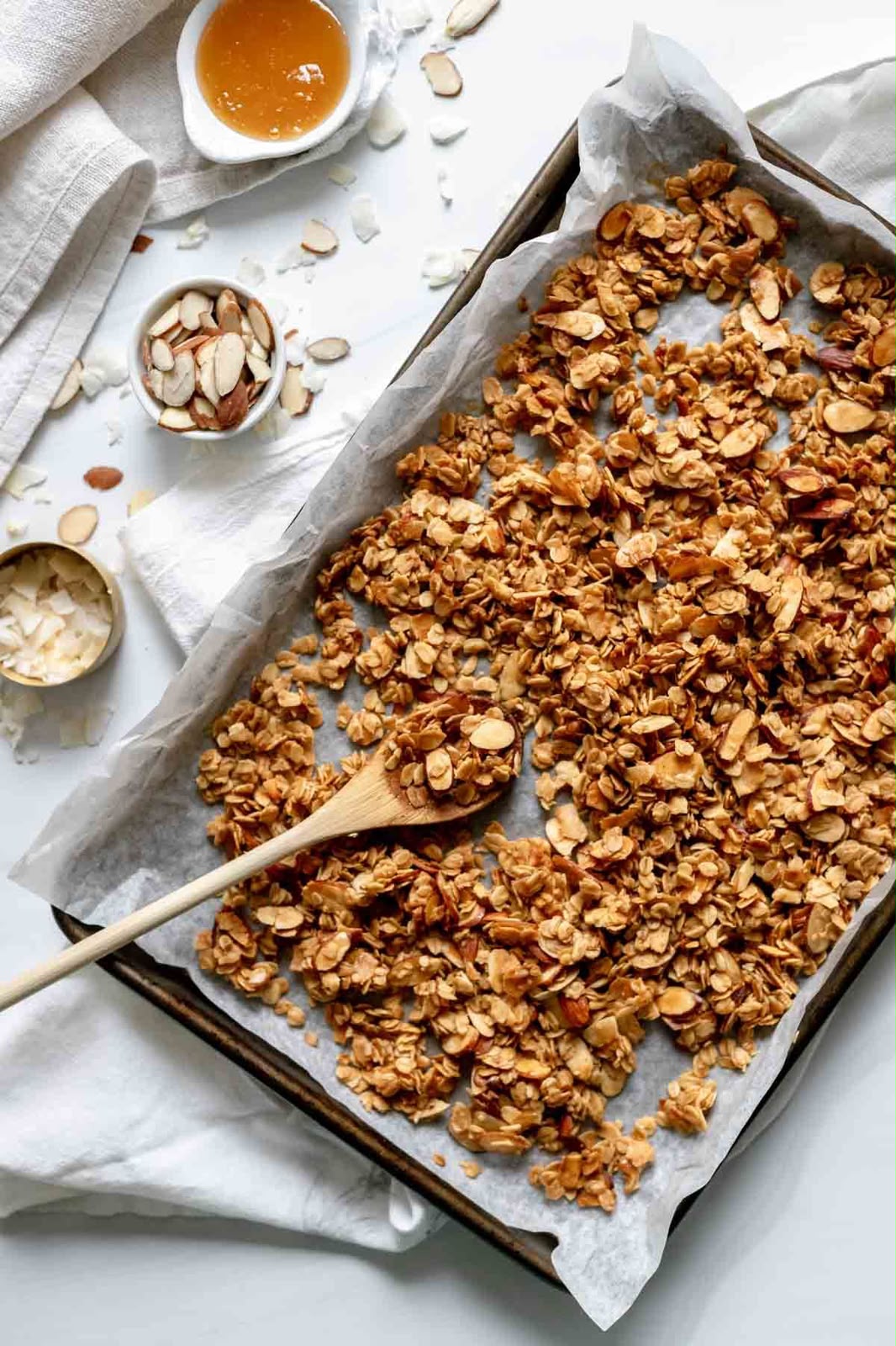 Granola spread across a parchment-lined baking sheet with a wooden spoon, styled with small bowls of sliced almonds, dried fruit, and cream on a white cloth background.