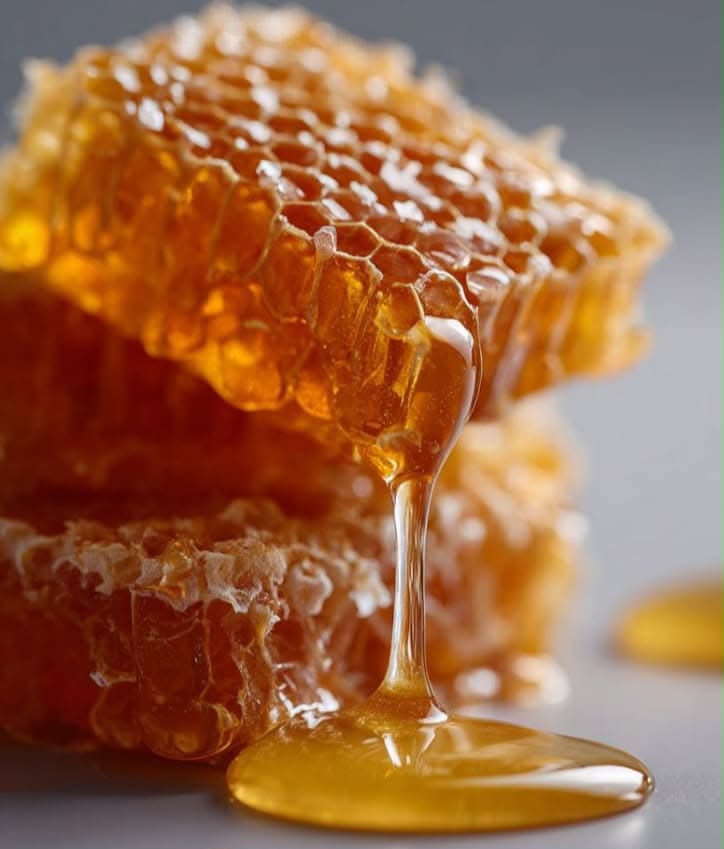 Close-up of honeycomb dripping with golden honey, with a glossy pool of honey in the foreground on a soft gray background.