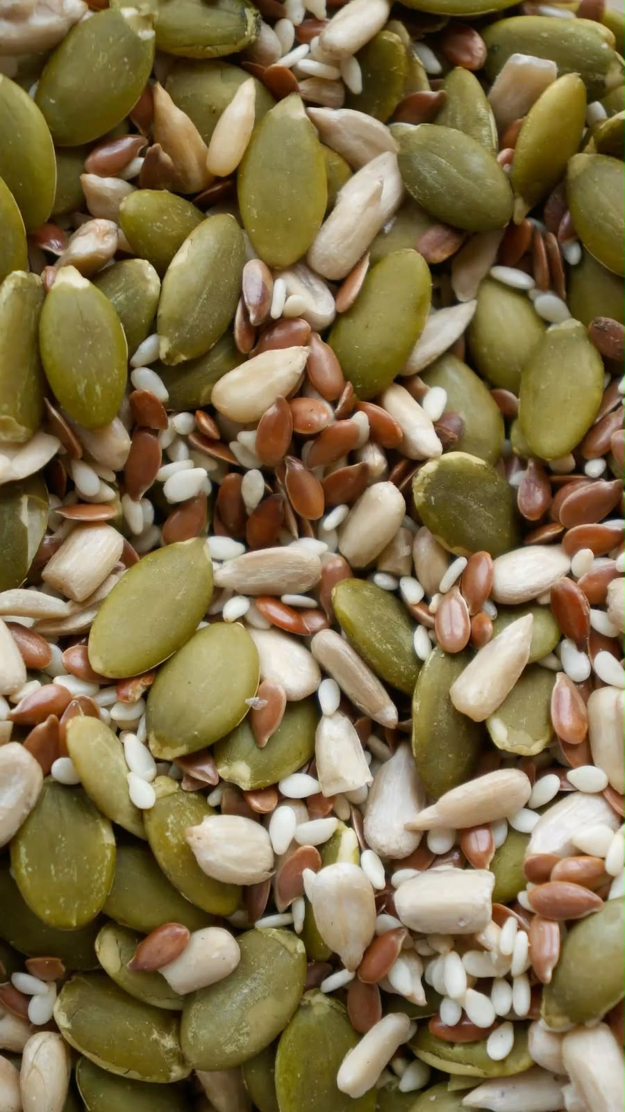 A close-up top-down view of mixed seeds including green pumpkin seeds, white sunflower kernels, and brown flaxseeds.